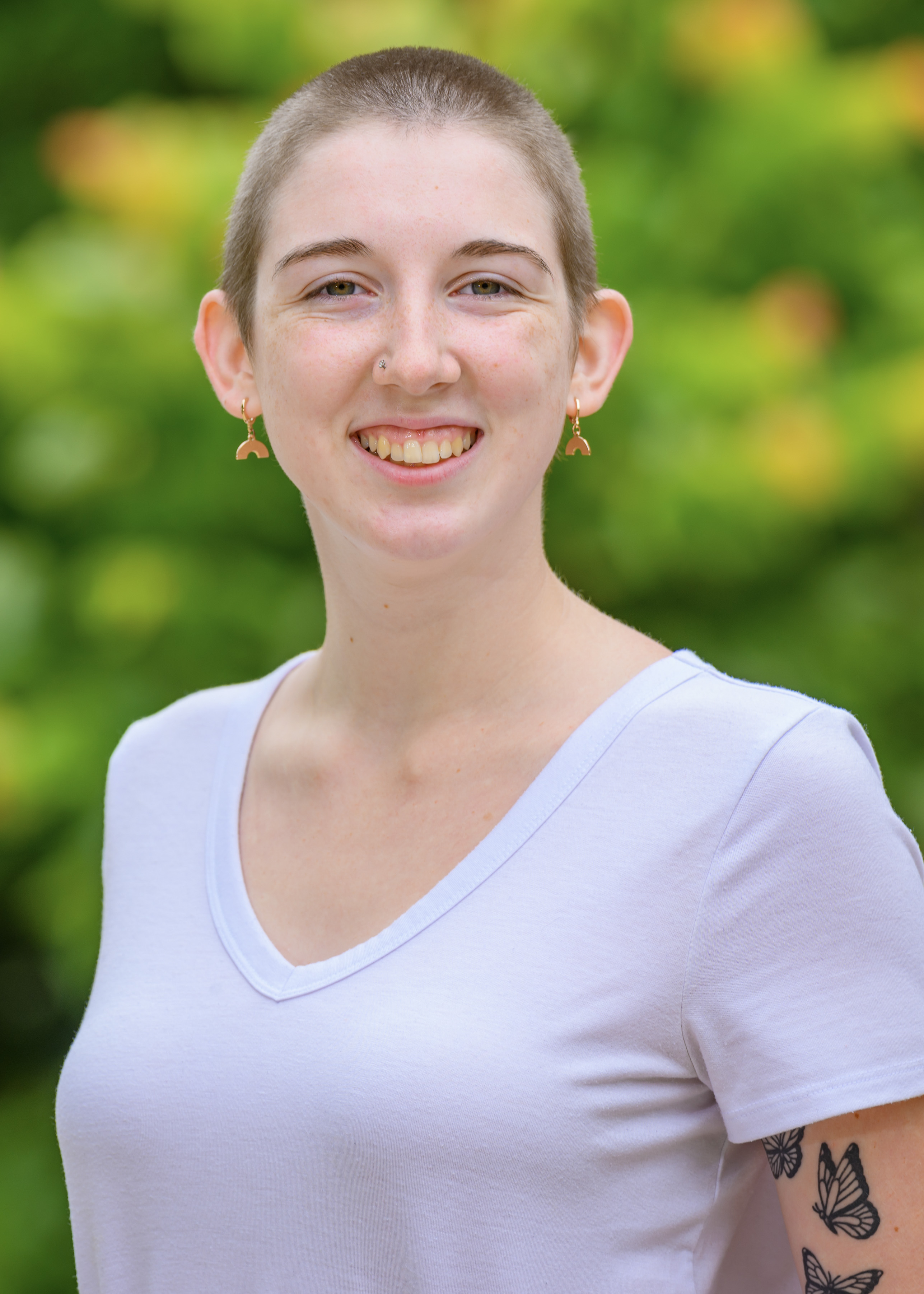 A young woman with very short hair, gold earrings, and a white shirt, facing forward and smiling.