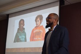 A Black man in a suit smiling in front of a projector screen.