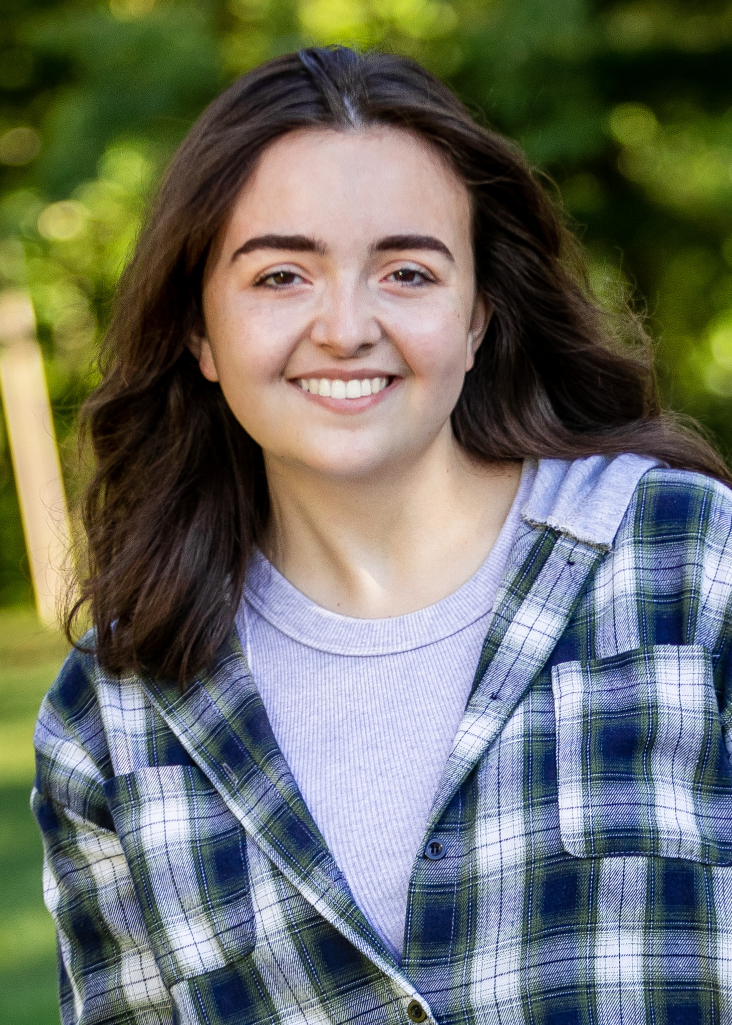 A woman with medium-length brown hair, wearing a gray shirt and a gray-and-blue jacket, facing forward and smiling.